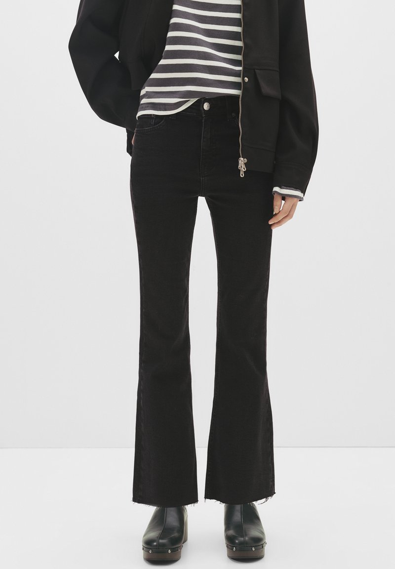 Osoba oblečená v černých rozšířených džínách, černých kotníkových botách se cvočky, pruhované košili a černé bundě na zip na jednoduchém pozadí.