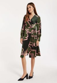 Wrap dress in green fabric with pink floral and geometric patterns. Features long sleeves, a tie waist, and a knee-length hem with a side slit.