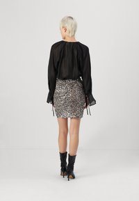 Crna prozirna bluzica dugih rukava s detaljima vezanja, uparena sa srebrnom mini suknjom od šljokica. Model nosi crne čizme na visoku petu. Glatke teksture kroz cijeli outfit.