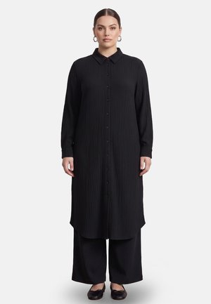 Vrouw draagt een lange zwarte knoopjes-overhemdjurk over een brede zwarte broek en zwarte schoenen, staand tegen een effen witte achtergrond.