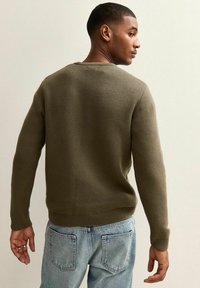 Maglione a maglia testurizzato verde oliva con maniche lunghe, polsini a coste e scollatura rotonda, indossato con jeans azzurri chiari.
