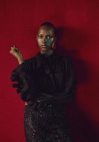 Blusa nera con maniche a volant, abbinata a una gonna nera con paillettes. Lo sfondo è di un rosso intenso. Il soggetto ha una pelle liscia e indossa orecchini.