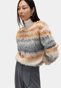 Weicher Pullover mit horizontalen Streifen in Beige, Orange und Grau. Rundhalsausschnitt, lockere Passform, strukturierte Oberfläche. Kombiniert mit hellgrauen Hosen.