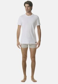 Mężczyzna stoi boso, ubrany w prostą białą koszulkę i szare majtki, na których brzegu znajduje się niewielkie czarne logo.