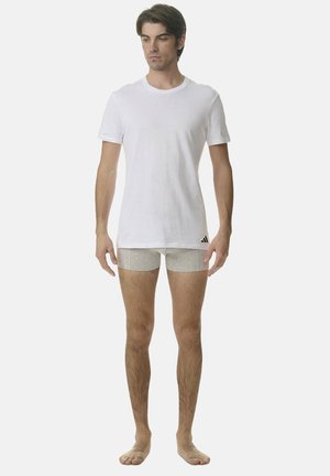 Mężczyzna stoi boso, ubrany w prostą białą koszulkę i szare majtki, na których brzegu znajduje się niewielkie czarne logo.