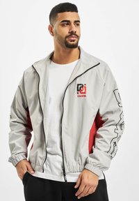 Light gray windbreaker jacket featuring a red panel, logo on the chest, zip front, elastic cuffs, and a casual fit.