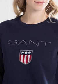 Námořnická modrá mikina s vyšitým nápisem "GANT" v světle šedé barvě a logem s erbem, hvězdami a pruhy v červené a bílé.