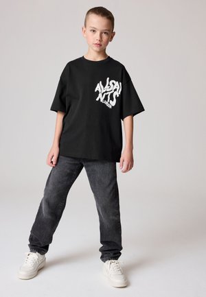 Băiat stând cu picioarele depărtate, purtând un tricou negru supradimensionat cu text alb, blugi gri închis și adidași albi pe un fundal simplu.