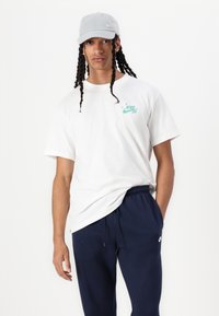 Camiseta branca de algodão com logótipo verde da Nike SB, combinada com calças de fato de treino azul-marinho. Modelo a usar um boné cinzento claro, com aba curva.