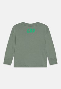 Majica dugih rukava u maslinasto zelenoj boji s okruglim izrezom, koja na leđima ima izvezeni zeleni logotip "GAP". Mekani materijal, standardni kroj, rukavi sa manžetama.