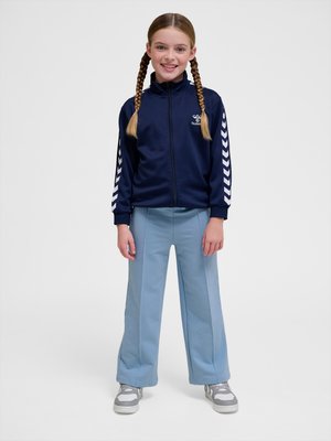 Smilende pige med fletninger iført en navy lynlåstrøje med hvide chevronstriber og lyseblå viddebukser.