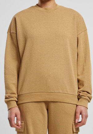 Osoba ubrana w luźną beżową bluzę i pasujące beżowe spodnie bojówki z bocznymi kieszeniami, stojąca ze zrelaksowanymi ramionami wzdłuż ciała.