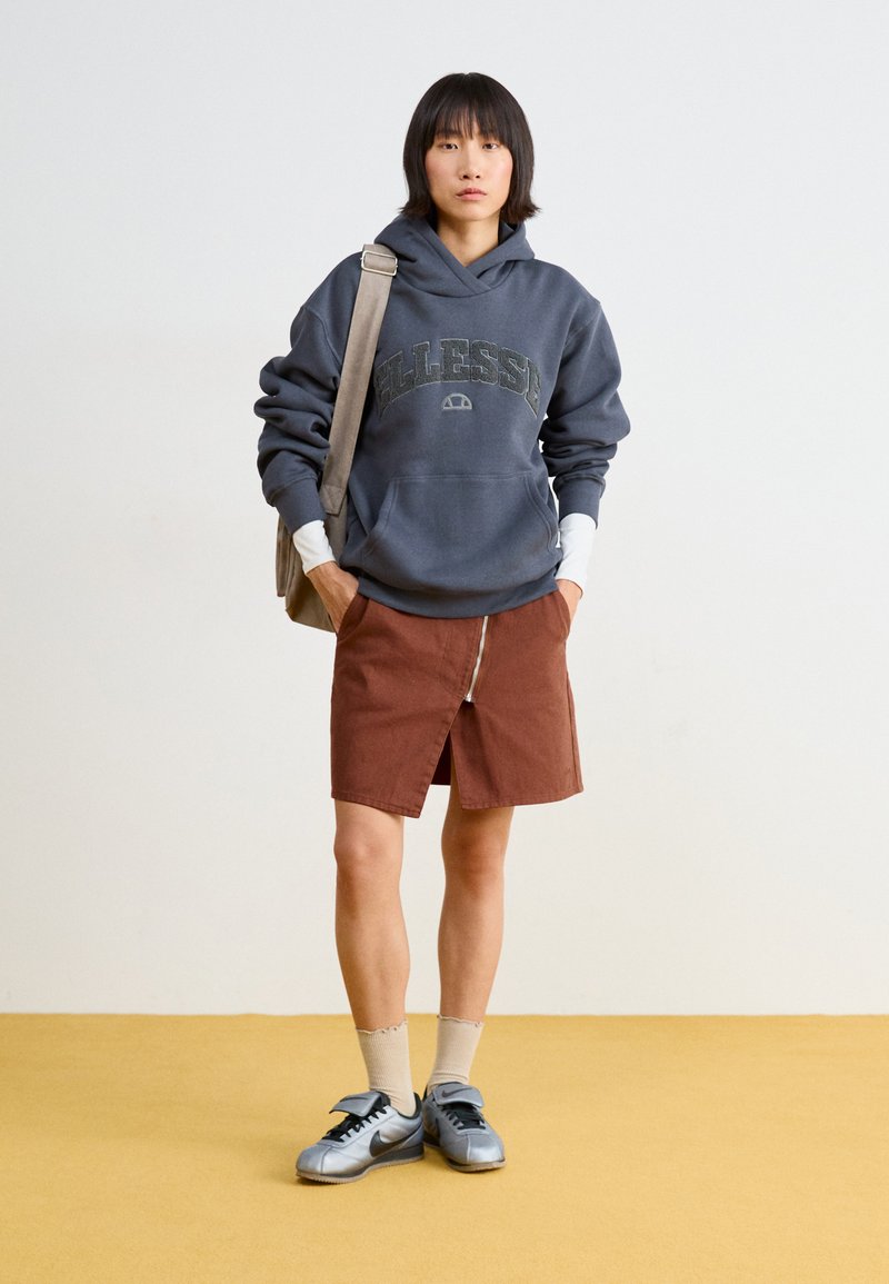 Sudadera con capucha de color gris oscuro con un logo, falda marrón con cremallera en la parte delantera, zapatillas grisáceas y calcetines tobilleros beige sobre un fondo de suelo amarillo.
