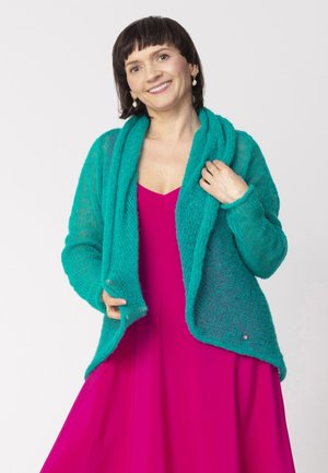 Kobieta uśmiechająca się, nosząca perłowe kolczyki, jasnoróżową sukienkę oraz turkusowy dziergany sweter, trzymająca brzegi swetra obiema rękami.