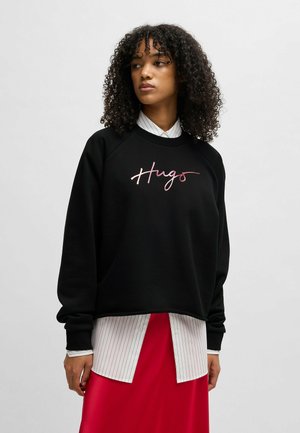 Zwarte sweatshirt met een cropped ontwerp, voorzien van roze geborduurde tekst "Hugs". Gelaagd over een wit gestreept overhemd met kraag en een rode rok.