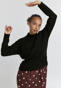 Schwarzer gestrickter Pullover mit hohem Kragen und fallenden Schultern, kombiniert mit einem kastanienbraunen Rock mit weißen Punkten.