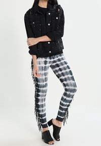 Veste en denim noir avec des détails déchirés et des clous argentés, associée à un legging tie-dye noir et blanc avec des franges. Mules noires.