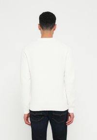 Uomo che indossa un maglione bianco a maniche lunghe e jeans blu scuro, in piedi di spalle davanti a uno sfondo bianco.