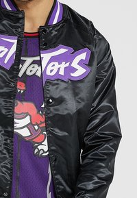 Chaqueta bomber de satén negra con un gráfico púrpura en el pecho, que presenta un cuello a rayas. Usada sobre una camiseta atlética púrpura con gráfico.