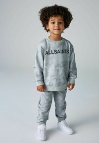 Pilka tinklelio spalvos džemperis su derančiomis kelnėmis ir laisvai krentantys rankogaliai, su juodu "ALLSAINTS" logotipu. Dėvima su baltais sportiniais bateliais.