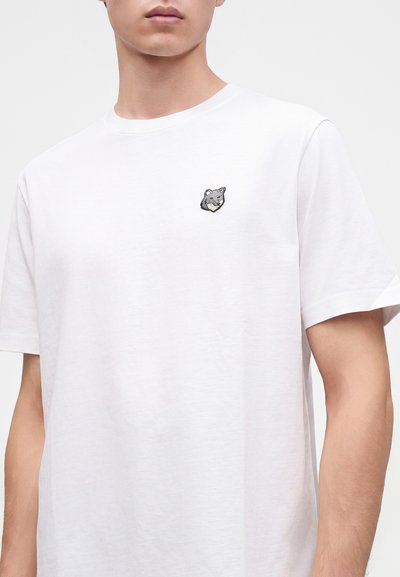 Νεαρό άτομο που φορά ένα απλό λευκό t-shirt με μικρή κεντημένη γκρίζα κεφαλή λύκου στο αριστερό στήθος, σε μονόχρωμο φόντο.