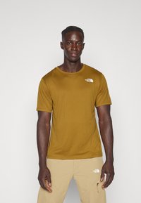 T-shirt de manga curta, amarelo mostarda, feito de um tecido suave, com um logótipo branco no lado esquerdo do peito. Modelo a usar calças bege.