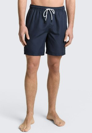 Mężczyzna w ciemnoniebieskich kąpielówkach ze białym sznurkiem, stojący boso na jasnym jednolitym tle.