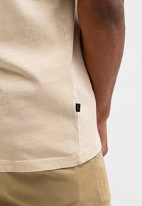 Beige katoenen T-shirt met een gestructureerd oppervlak en zijkleurlabel. Korte mouwen, met een rechte zoom en minimale stikseldetails.