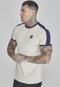 Camiseta crema con acentos en azul marino en los hombros, diseño ajustado y un pequeño logo negro en el lado izquierdo del pecho. Tela suave, mangas cortas.