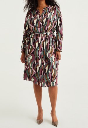 Vrouw draagt een knielange, lange mouwen jurk met een multicolor abstract motief en een zelfgeknoopt riempje, gecombineerd met bruine puntige hakken.