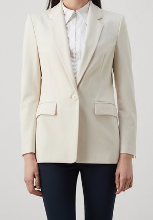 Kvinde iført en tætsiddende cremefarvet blazer over en hvid skjorte med knapper og mørke bukser, stående mod en enkel lys baggrund.
