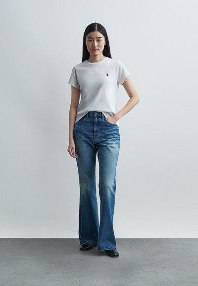 Νεαρή γυναίκα που στέκεται με το ένα χέρι στην τσέπη, φοράει λευκό κοντομάνικο πουκάμισο με λογότυπο και μπλε τζιν παντελόνι με καμπάνα, μπροστά από απλό φόντο.