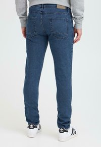 Kitsas lõikega tumed blue denim teksad, millel on õmmeldud tagataskud ja nahkplaat vööl, kantud valgete kingadega.