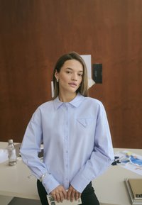 Chemise boutonnée bleu clair avec un détail de poche, manches longues, et col, portée sur un pantalon noir. Texture de tissu lisse et net.