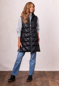 Woman wearing long black quilted vest, gray sweater, blue jeans, and black boots standing on wooden floor against plain background.