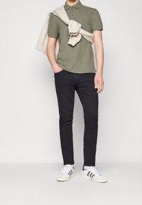 Olivově zelená polokošile se třemi knoflíky, spárovaná s černými slim-fit džíny a bílými teniskami se šedými pruhy; světlý svetr přehozený přes ramena.