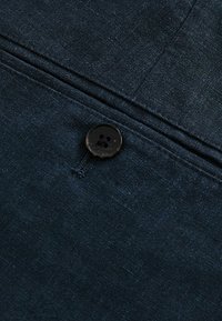 Trezire din in de culoare albastru închis, cu o textură netedă, având un nasture negru cu patru găuri. Include un detaliu discret de cusătură.