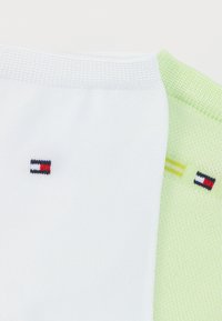 Δύο κάλτσες τοποθετημένες δίπλα-δίπλα: η μία λευκή με ένα μικρό λογότυπο σε σκούρο μπλε, κόκκινο και λευκό· η άλλη ανοιχτοπράσινη με μια κίτρινη ρίγα και το ίδιο λογότυπο.