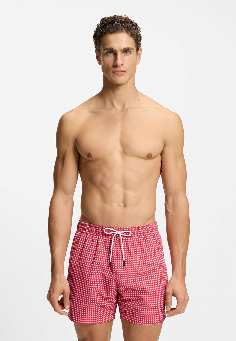 Homme musclé debout, portant un short de bain rouge et blanc à motif avec un cordon blanc, fond blanc uni.