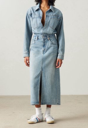 Person iført en lyseblå denimskjorte og matchende højtaljet midinederdel med frontslids, hvide sokker og hvide sneakers med blå og røde striber.