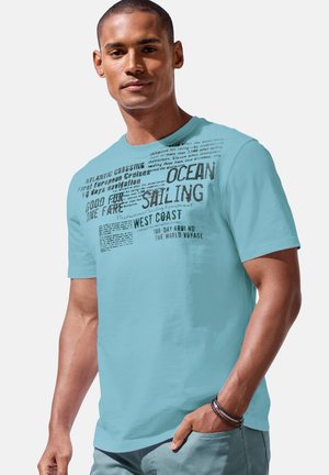 Mann trägt hellblaues T-Shirt mit schwarzem segelthemenbezogenem Text, steht mit einer Hand in der Tasche vor einfarbigem Hintergrund.