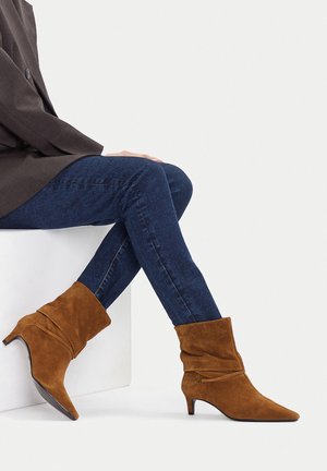 Osoba sedící se zkříženýma nohama, oblečená v tmavě modrých úzkých džínách, hnědých semišových kotníkových botách na středním podpatku a tmavém saku.