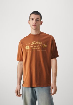 Camiseta de algodón marrón con el logo de "New Era Cap Co." en dorado, de manga corta, con cuello redondo y un ajuste relajado, combinada con jeans de color azul claro.
