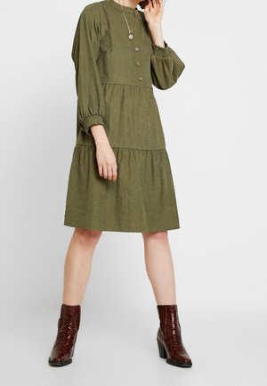 Olivgrünes Kleid mit geknöpftem Vorderteil, Ballonärmeln und gestufter Rock. Mit gerafften Manschetten und strukturierter Stoff, kombiniert mit Stiefeletten.