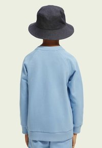 Felpa azzurra chiara con polsini e orlo a costine, abbinata a un cappello a secchiello grigio scuro. La vista mostra la parte posteriore dell'outfit.