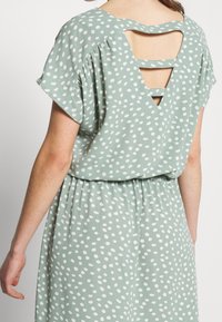 Femme portant une robe vert clair avec des pois blancs irréguliers, présentant des manches courtes et un dos découpé avec des bretelles horizontales.