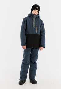 Blauwe en zwarte waterdichte jas met logo en ritszak, gecombineerd met bijpassende broek en zwarte winterlaarzen. De outfit is geschikt voor koud weer.