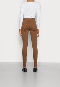 Jeans ajustados de talle alto color marrón, hechos de una mezcla de algodón, con bolsillos traseros y una textura suave, combinados con una blusa de manga larga blanca.
