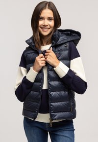 Navy nieprzemakalna kamizelka puchowa z kapturem, zapinana na zamek, o wyściełanym fasonie. Noszona na pasiakowy sweter; zawiera zamki oraz akcent z logo.