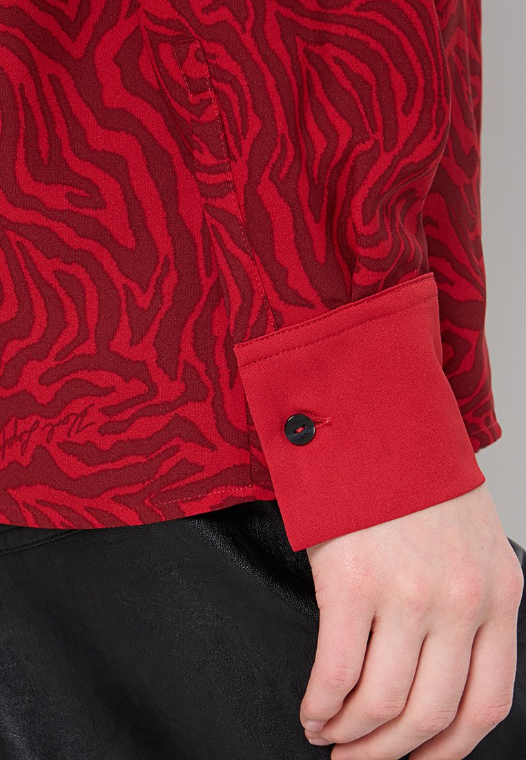 Chemise rouge avec un motif rayé zèbre texturé, présentant un poignet rouge uni avec un détail de bouton noir. Tissu lisse avec un léger éclat.